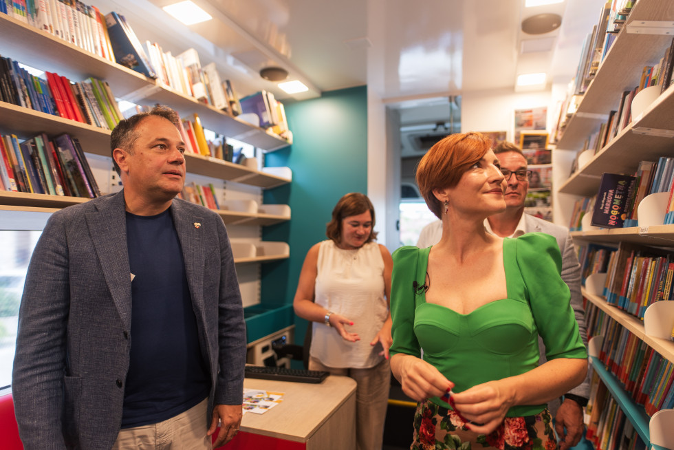 Ministra, župan in direktorica stojijo v bibliobusu in si ogledujejo knjige na policah.