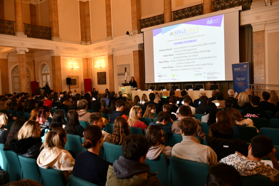 Pogled na govornico, kjer stoji državna sekretarka Humar, in mnogo mladih gledalcev.