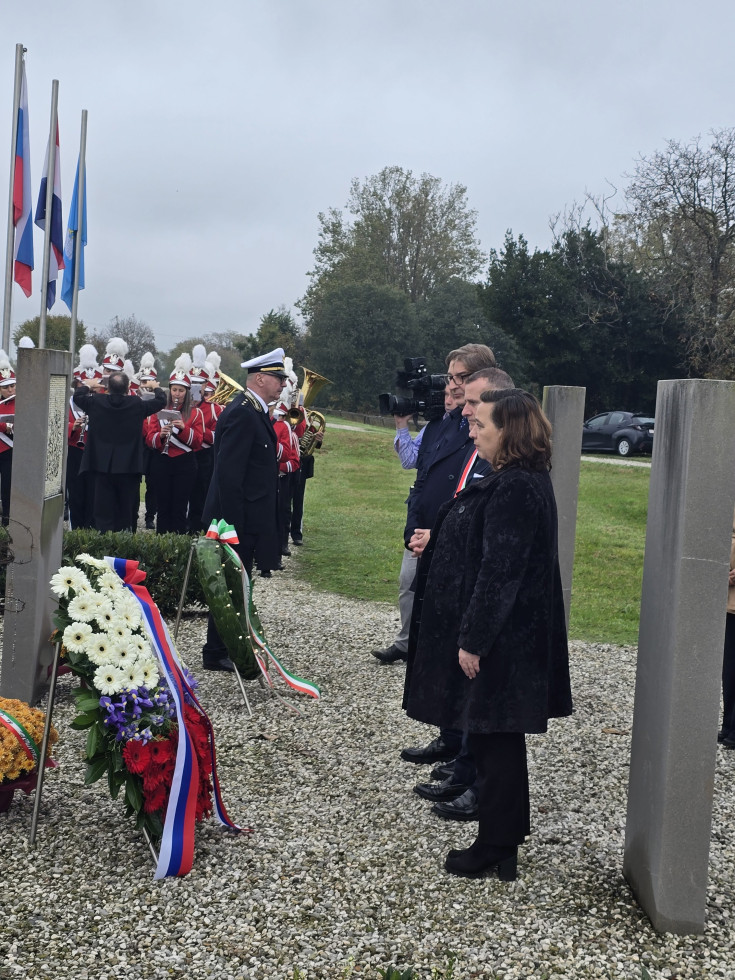 Državna sekretarka in generalni konzul pred spomenikom z vencem, v ozadju godba.