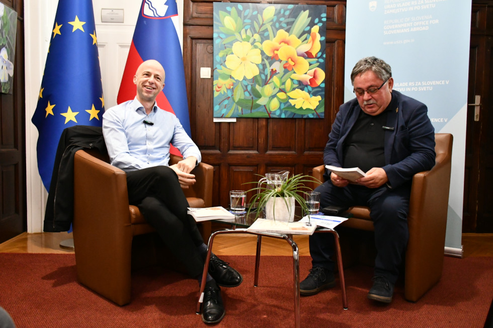 dr. Tomaž Simetinger in dr. Boris Jesih med pogovorom za mizo, v ozadju zastava in transparent urada, na mizi in v rokah Jesiha knjige.