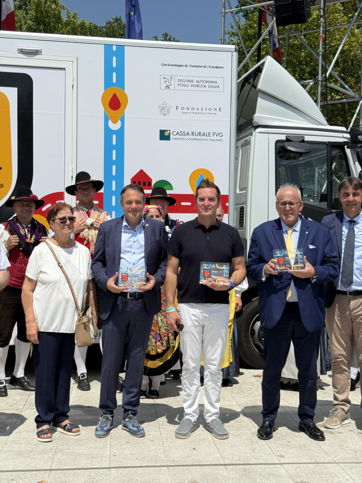 Alt text: "Minister Arčon, župan Nove Gorice in župan Gorice stojijo v ospredju pred prireditvenim vozilom, v rokah držijo spominske plakete; za njimi so člani folklorne skupine v tradicionalnih nošah."
