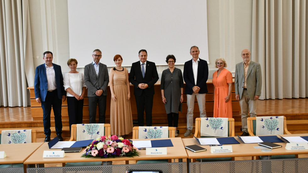 Alt text: "Minister Arčon in ministrica Vrečko stojita skupaj s predstavniki kulturnih krovnih organizacij v dvorani pred odrom, za mizo z razporejenimi mapami in šopkom rož, pripravljeno za podpis uradnih dokumentov."