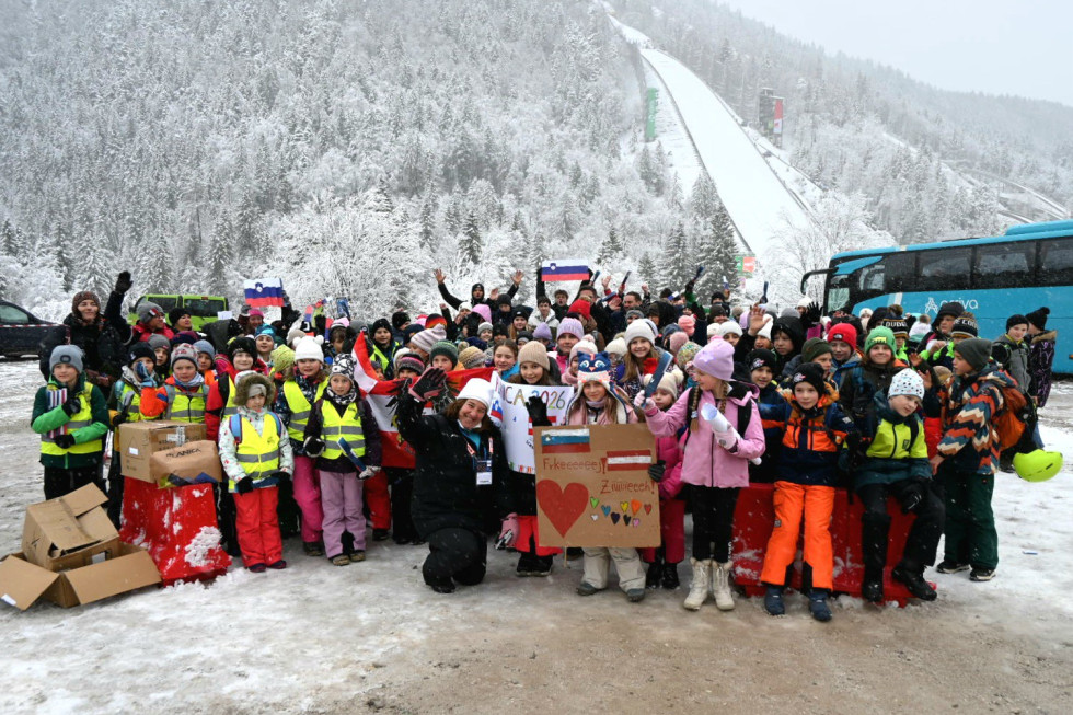 Skupinska slika z otroci z zastavami, plakati, v ozadju letalnica.