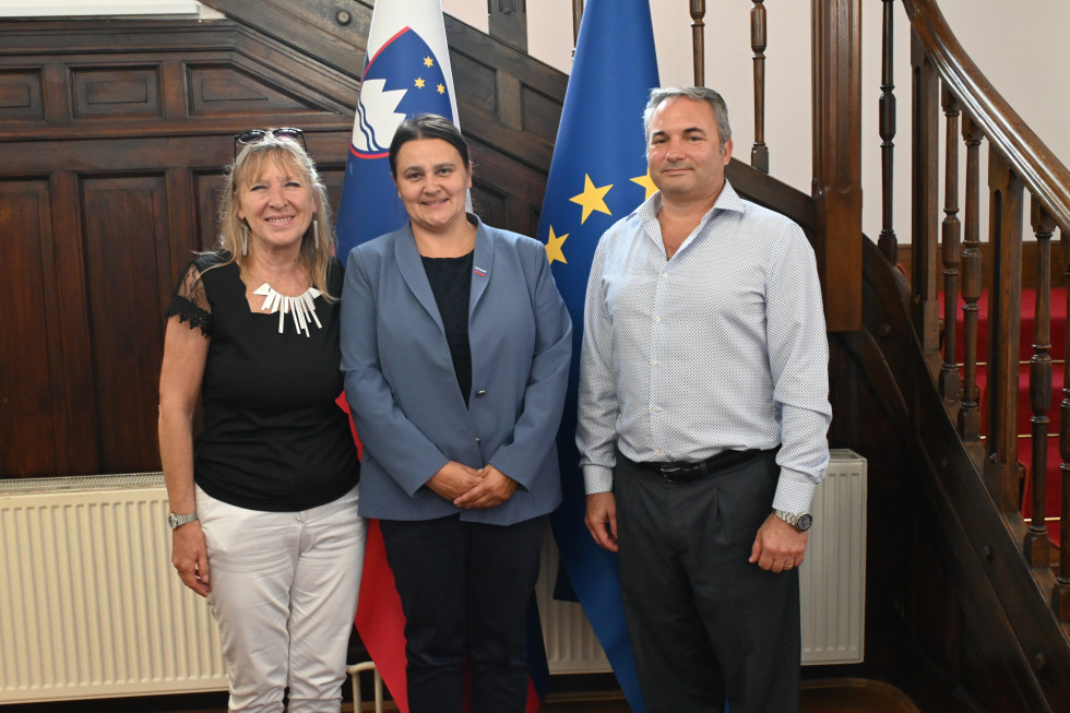 Mariana Pirc, Suzana Lep Šimenko in Damijan Ahlin pred slovensko in evropsko zastavo v avli urada.
