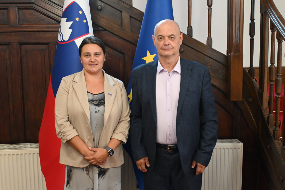 Ministrica in gospod Jovičič pred zastavami v avli urada.