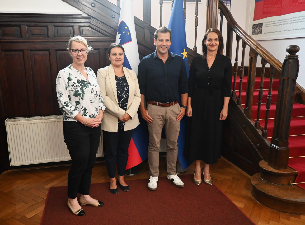 Ministrica in državna sekretarka z gosti pred zastavami v avli urada.