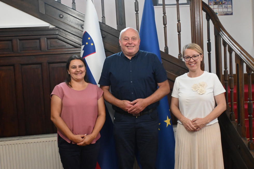 Ministrica in državna sekretarka z gostom pred zastavami v avli urada.