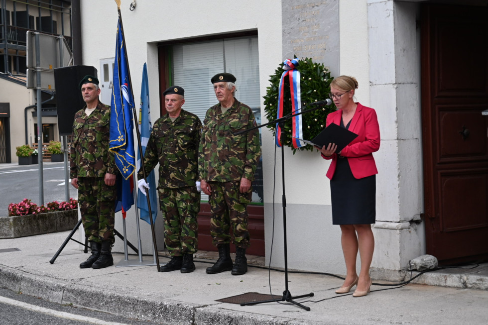Državna sekretarka med nagovorom, za njo venec na spominski plošči, na levi vojaki in zastave.
