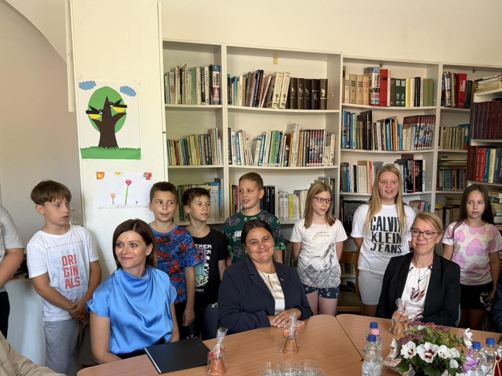 Tri ženske sedijo za mizo, otroci stojijo za njimi. Zadaj knjižne police s knjigami.