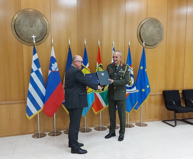 Veleposlanik Matjaž Longar in generalmajor Dimitrios Choupis si izmenjujeta sporazuma v ozadju zastave